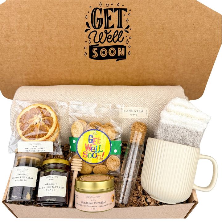 An open gift basket box with the text 'GET WELL SOON' featuring a variety of get well gifts including a beige throw blanket, a striped ceramic mug, assorted tea sets, a jar of honey, dried orange slices, cookies, a hand-poured soy candle, and a wooden honey dipper.