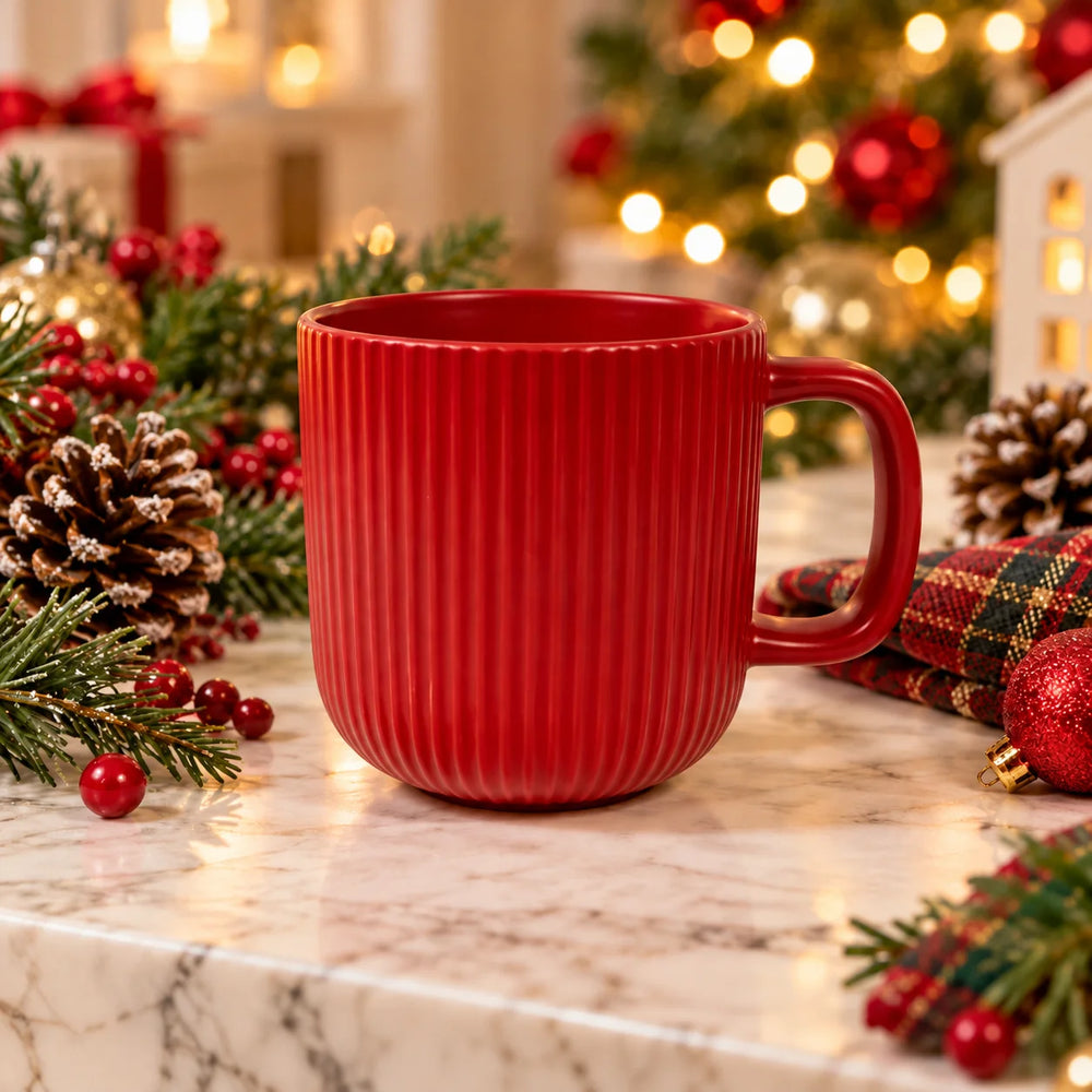 A red ribbed ceramic mug with a handle, set on a marble surface surrounded by Christmas decorations including pine branches, red berries, pinecones, and a plaid napkin, with a blurred Christmas tree in the background.