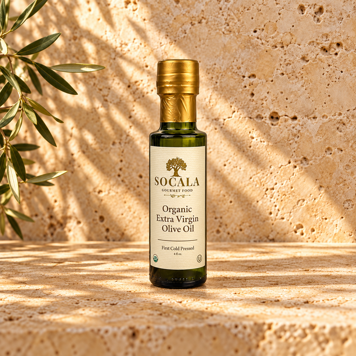 A bottle of Socala Organic Extra Virgin Olive Oil stands on a stone surface with warm sunlight and shadow patterns, next to olive branches.