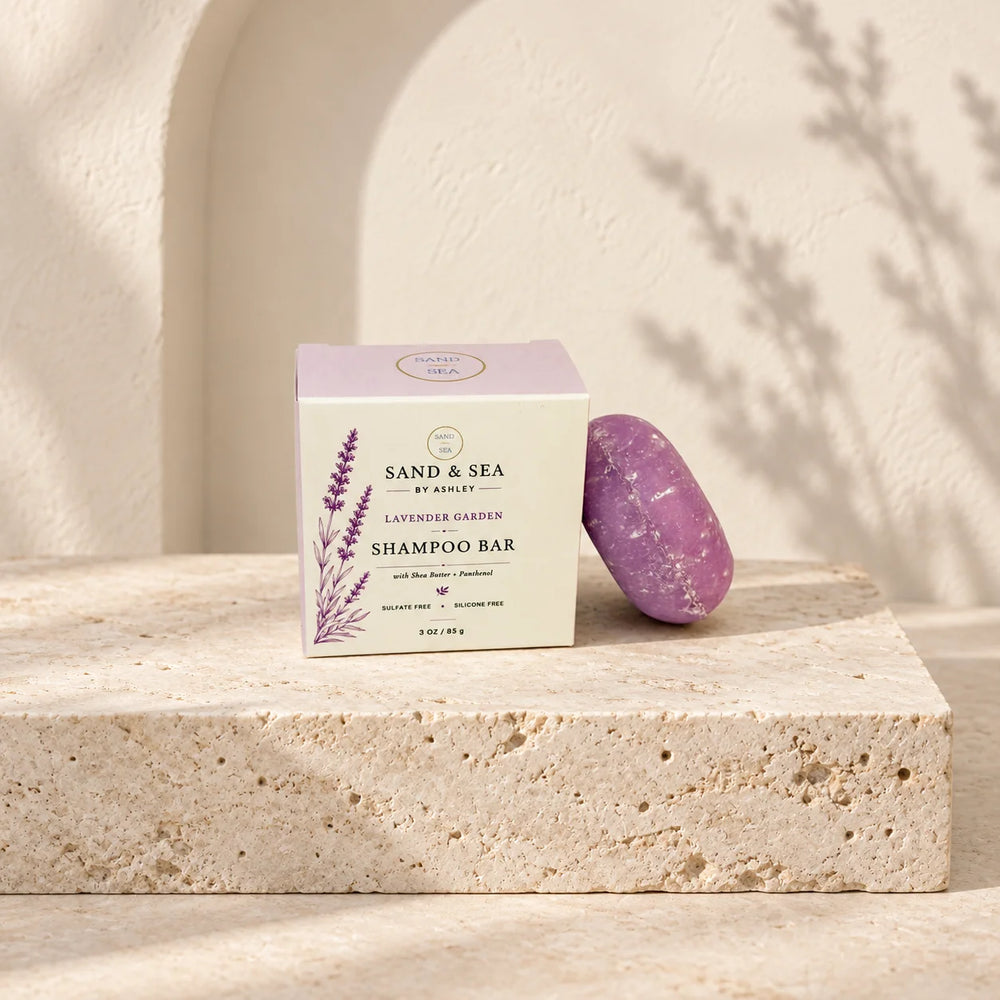 A boxed shampoo bar and a purple oval bar are displayed on a light stone pedestal against a warm beige wall with soft shadows. The box reads 'Sand & Sea by Aisley' and 'Lavender Garden Shampoo Bar.