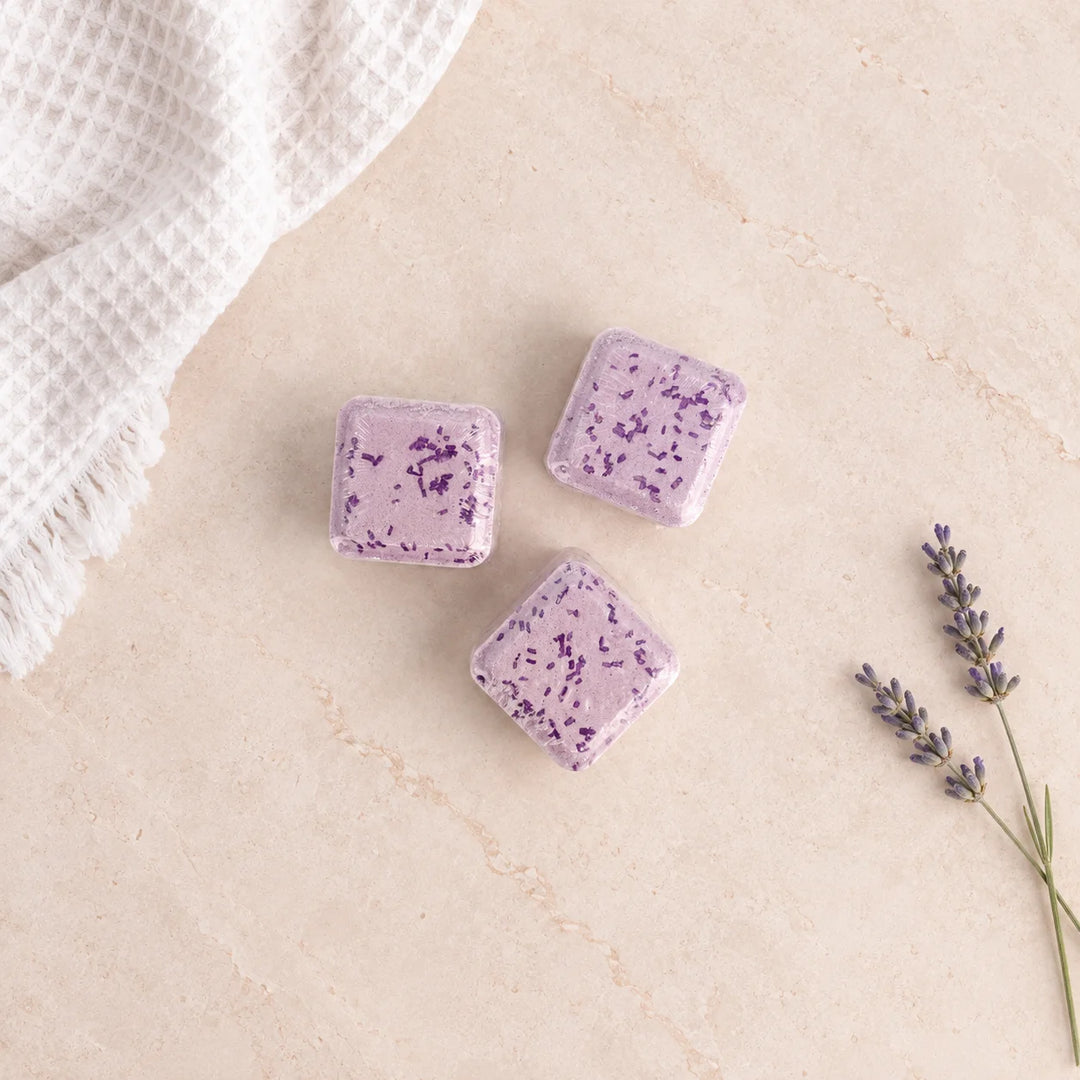 Three lavender bath cubes with purple botanical flecks, arranged on a beige stone surface next to a white textured cloth and a sprig of lavender.