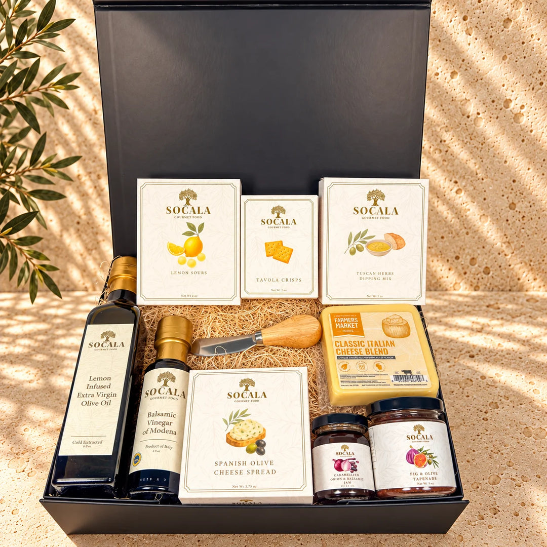 A Socala gourmet gift hamper set in an open black box, featuring lemon olive oil, balsamic vinegar, Spanish olive cheese spread, crackers, cheese, and fruit preserves, arranged on a textured beige surface with dappled sunlight and olive branches.
