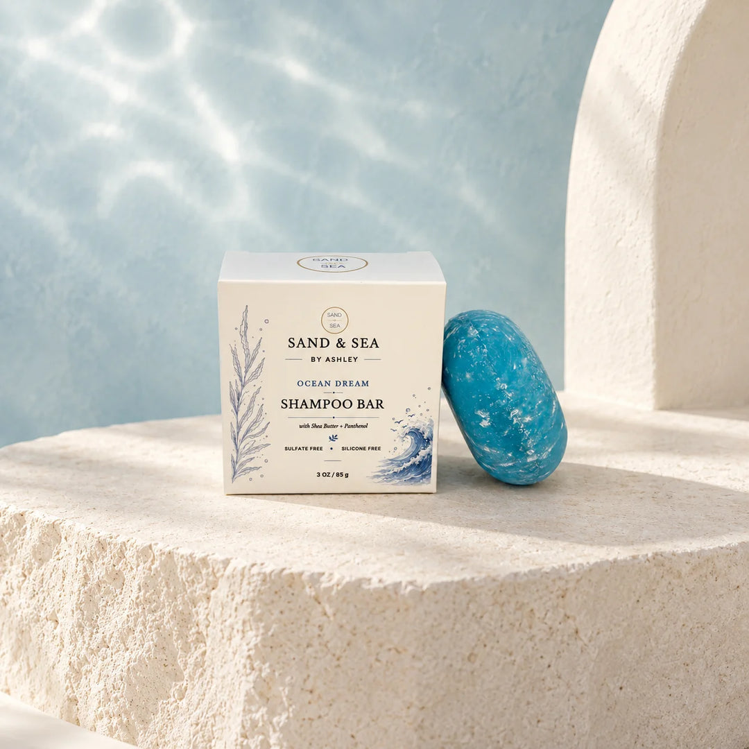 A boxed shampoo bar and a blue stone bar are displayed on a textured stone surface with a soft blue water-like backdrop. The box reads Sand &amp; Sea by Ashley Ocean Dream Shampoo Bar.