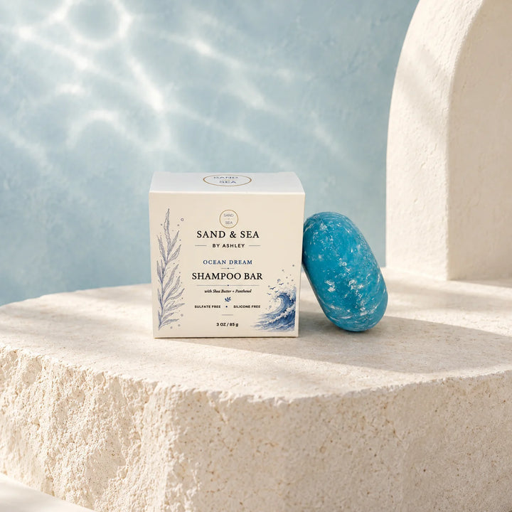A boxed shampoo bar and a blue stone bar are displayed on a textured stone surface with a soft blue water-like backdrop. The box reads Sand &amp; Sea by Ashley Ocean Dream Shampoo Bar.