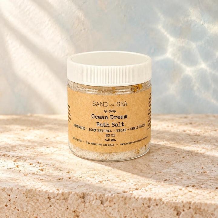 A small clear plastic jar of Sand + Sea by sandy Ocean Dream Bath Salt sits on a textured stone surface with a softly blurred water-like background. The jar has a white screw-top lid and a tan paper label with black text.