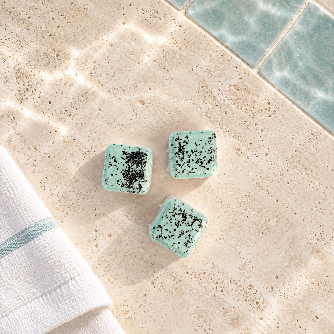 Three small square bath salts in a light mint green color with black speckles, arranged on a beige stone surface with a folded white towel and blue tiles in the background.