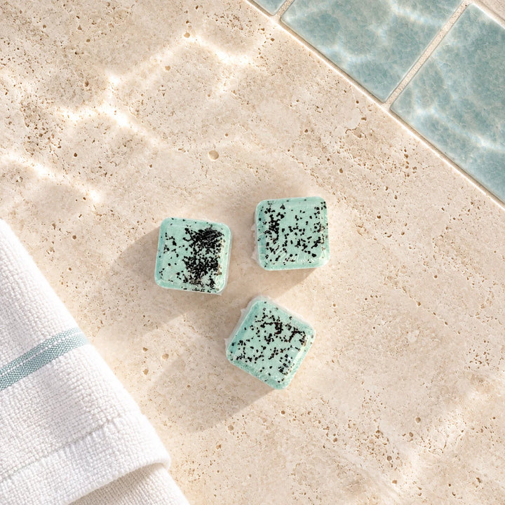 Three small square bath salts in a light mint green color with black speckles, arranged on a beige stone surface with a folded white towel and blue tiles in the background.