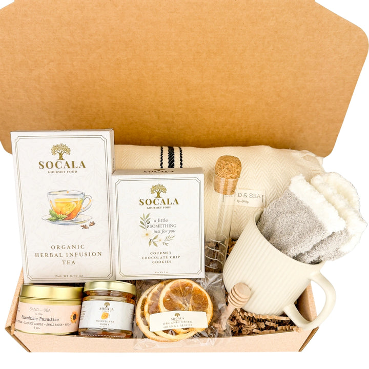Blanket and tea gift set with SOCALA tea bags, a mug, and various tea-related items in an open cardboard box.