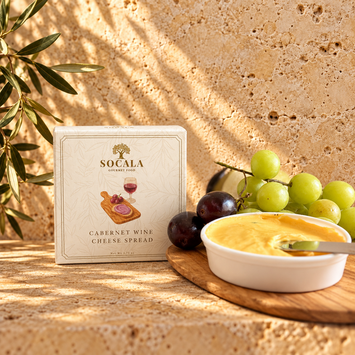 A boxed food product labeled SOCALA Cabernet Wine Cheese Spread stands on a stone surface beside a white bowl of yellow cheese spread, green and purple grapes, and a leafy plant against a textured beige wall with dappled sunlight.
