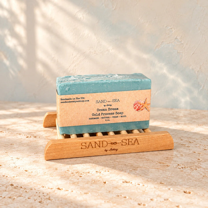 A rectangular bar of Sand by Sea Ocean Dream Gold Process Soap in a muted teal-blue color, resting on a wooden soap dish. The soap has a kraft paper label with a starfish graphic.