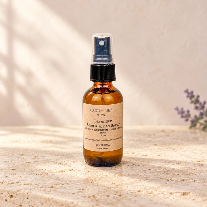 An amber glass spray bottle of Sand of Sea by Daisy Lavender Rose &amp; Tansy Spray, standing on a textured beige stone surface with soft shadow overlays and a small purple flower sprig in the background.