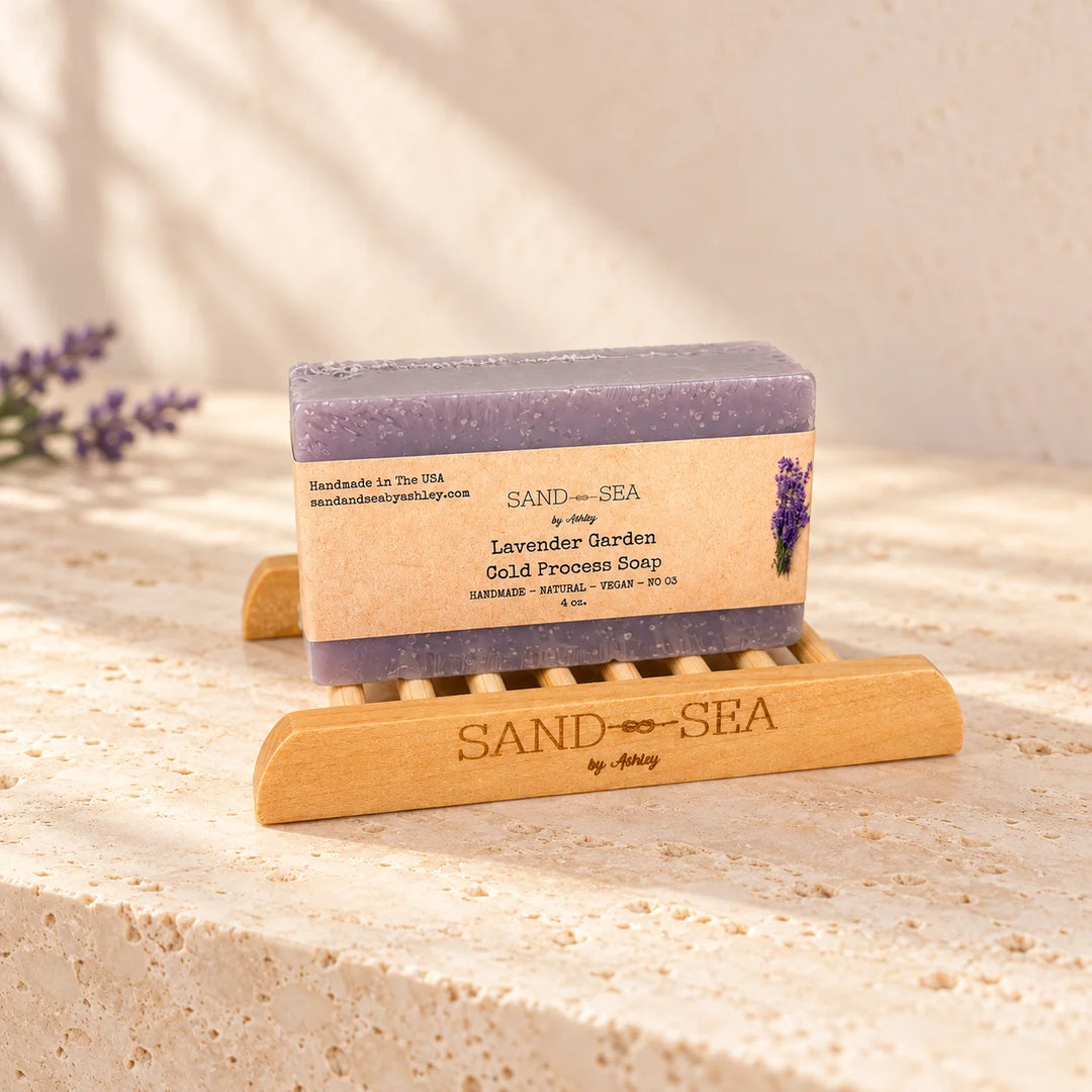 A bar of lavender garden gold process soap by Sand Sea, resting on a wooden soap dish on a textured stone surface with soft shadows.
