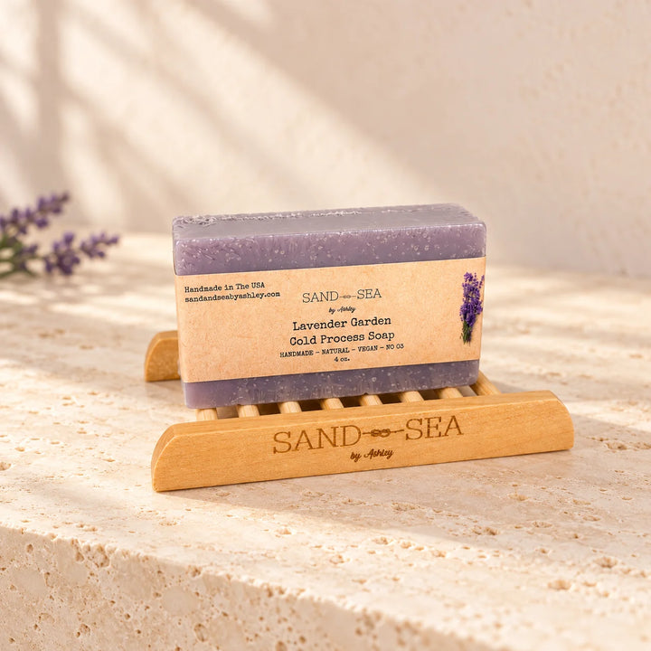 A bar of lavender garden gold process soap by Sand Sea, resting on a wooden soap dish on a textured stone surface with soft shadows.