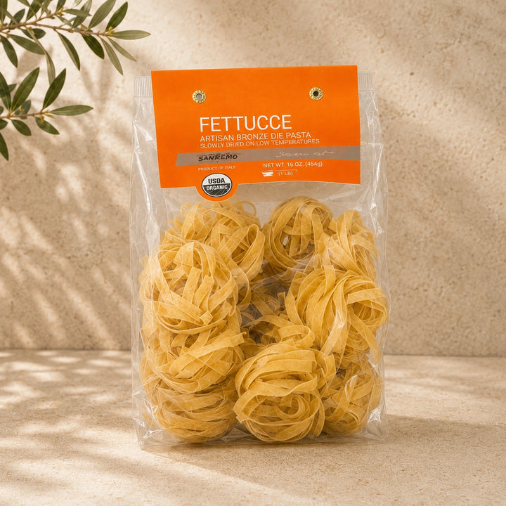 A clear plastic bag of Fettuccine Artisan Bronze Die pasta with an orange label, standing on a beige stone-like surface with leaf shadows and olive branches in the background.