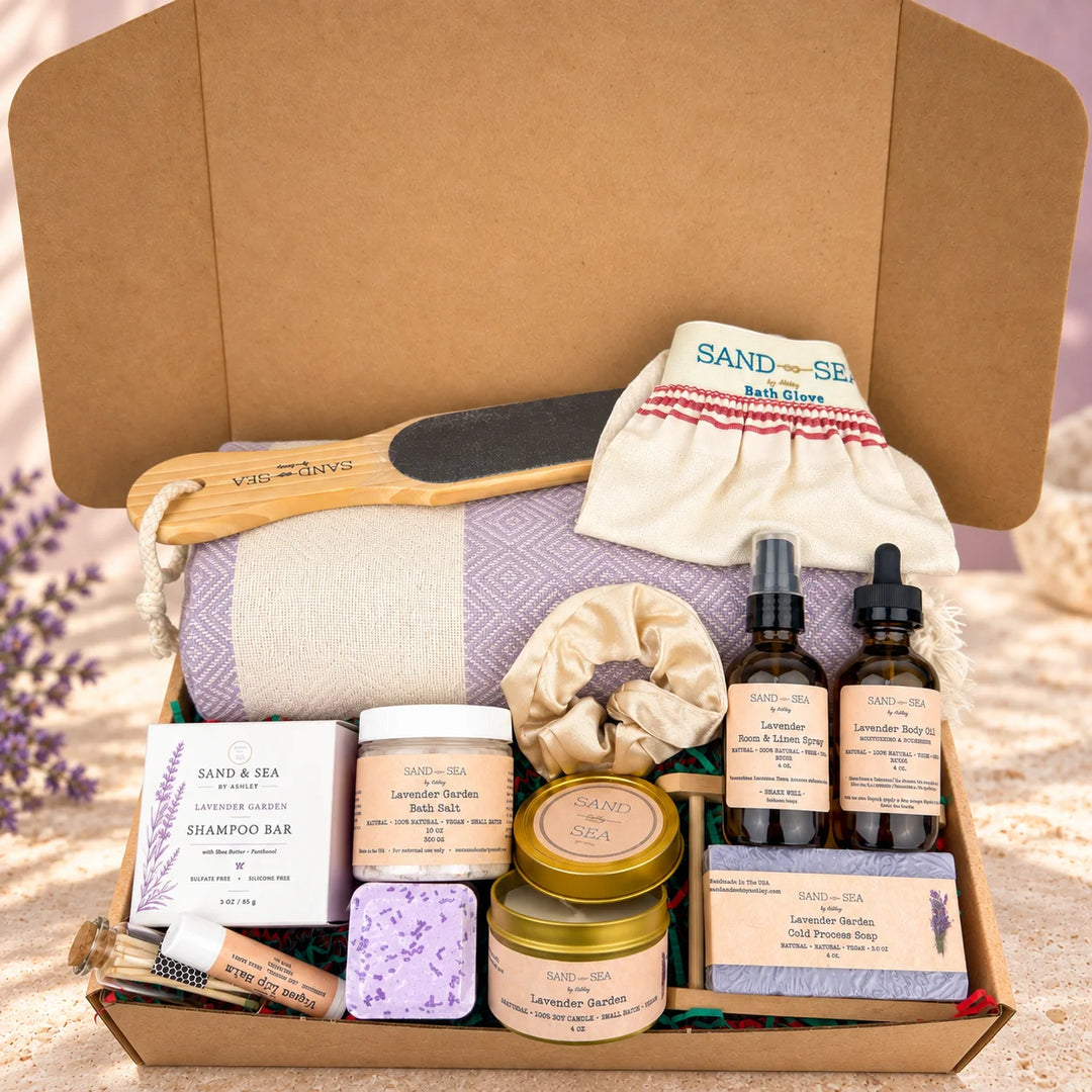 A curated bath and body gift set arranged inside an open kraft cardboard box. Visible items include a lavender-themed bath glass, a wooden scrubber, a cream towel, lavender body wash, lavender bath salts, lavender shampoo bar, lavender soap, lavender body butter, lavender foot soak, and amber dropper bottles.