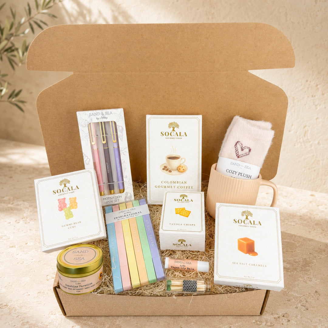 A curated gift box set from Socala, featuring coffee, sweets, stationery, and a plush toy arranged inside an open brown cardboard box with shredded paper. Visible items include Colombian Quadruple Coffee, sea salt caramel candy, pastel pencils, a notebook, a candle tin, and a pink plush toy.