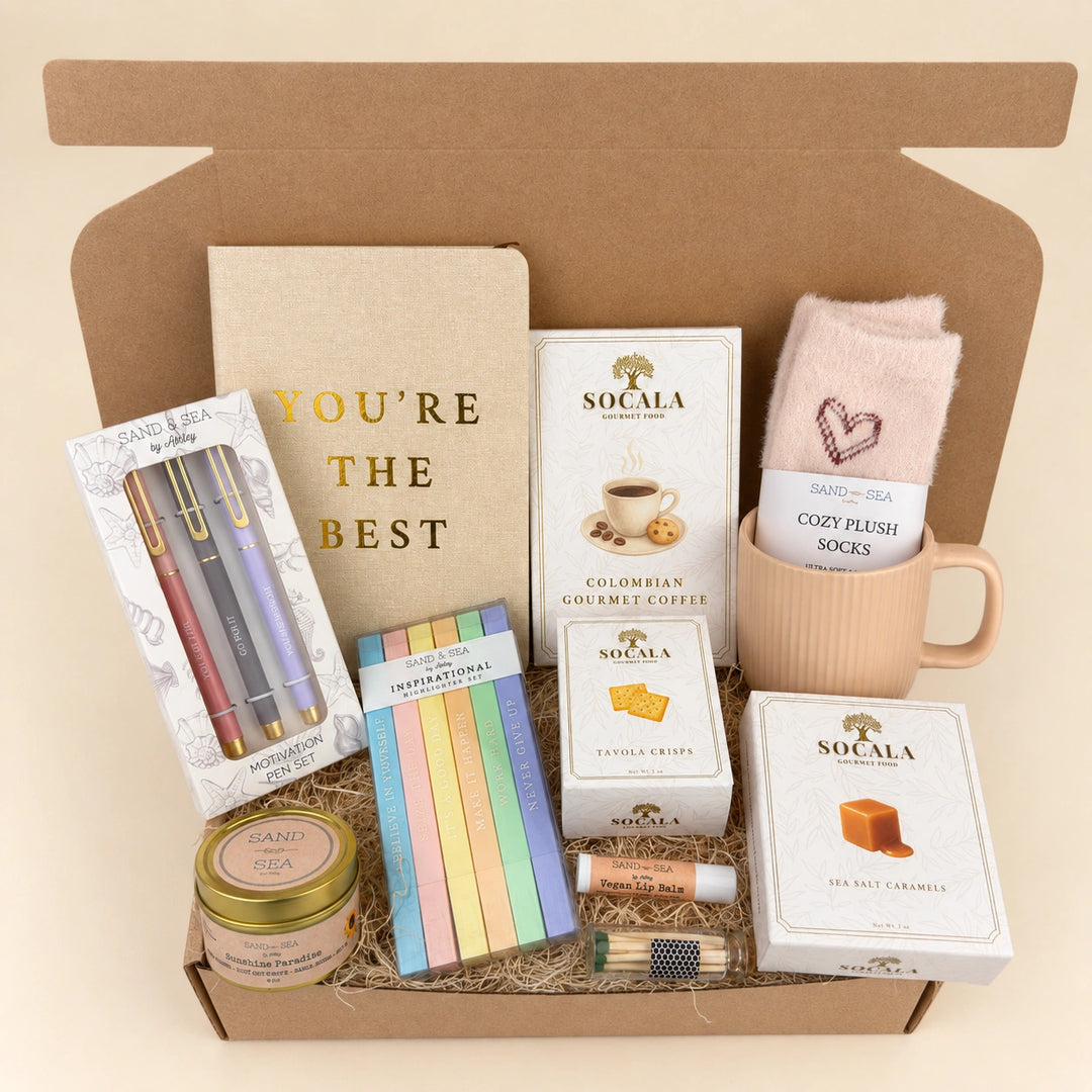 A curated gift box set in an open brown cardboard box, featuring a notebook that reads 'YOU'RE THE BEST', Socala coffee boxes, pastel sticky notes, a candle tin, pens, a lip balm tube, and plush socks in a pink mug.