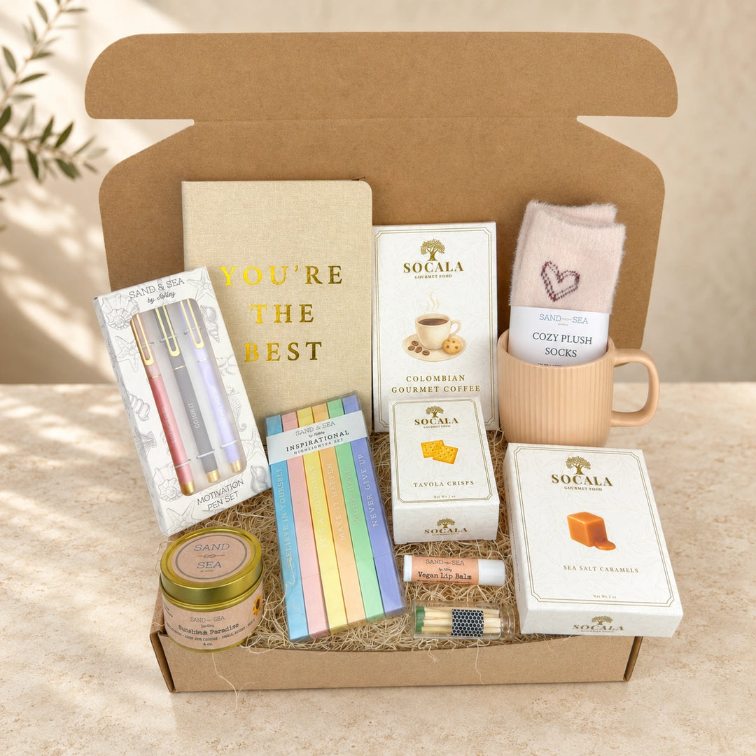 A curated gift box set in an open brown cardboard box, featuring a notebook that reads 'YOU'RE THE BEST', Socala coffee boxes, pastel sticky notes, a candle tin, pens, a lip balm tube, and plush socks in a pink mug.