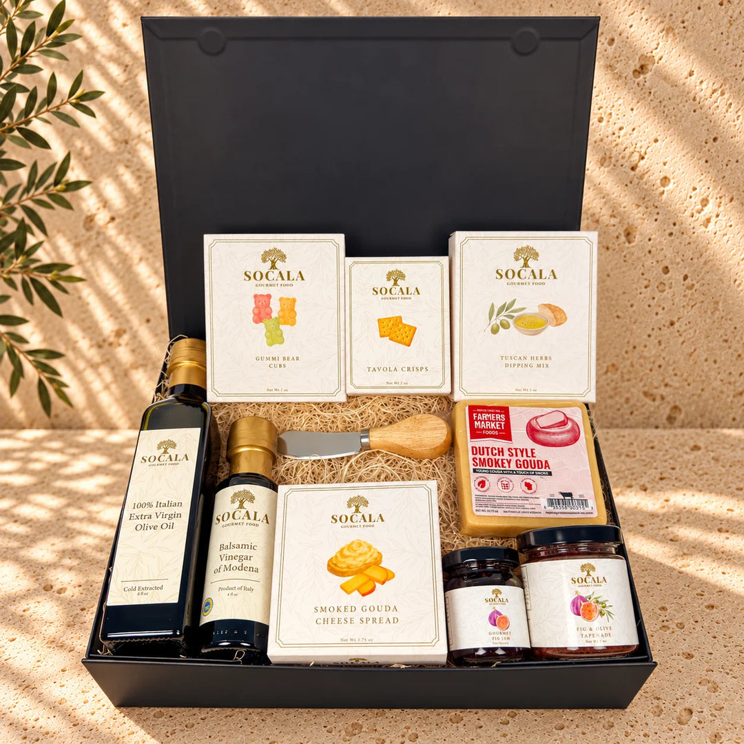 A gourmet gift hamper from Socala, presented in an open black box filled with cheese, spreads, olive oil, vinegar, and crackers, arranged on a textured beige stone surface with warm sunlight and shadows.