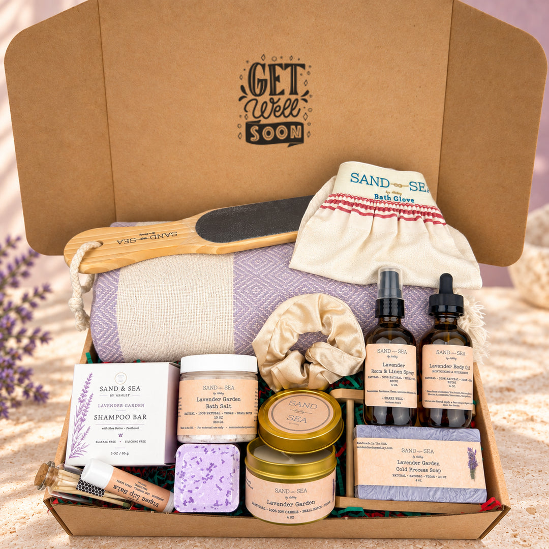 A curated bath and body gift box from Sand & Sea, filled with lavender-themed products including a lavender shampoo bar, lavender body wash, lavender bath salts, lavender soap, two amber dropper bottles, a lavender scrunchie, a lavender bath glove, a wooden massage tool, and a lavender pouch, all arranged in an open kraft cardboard box with the brand name printed inside.