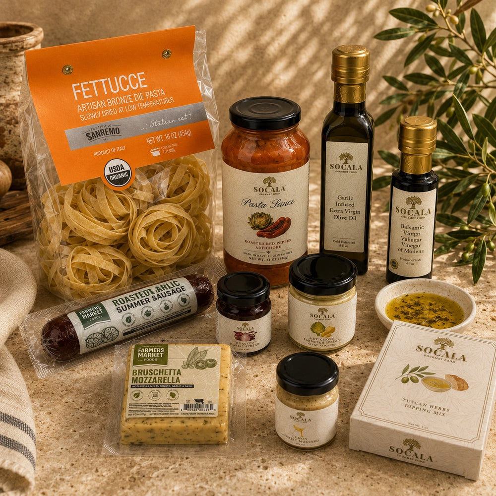 A styled flat-lay of a Sogala family dining set on a beige stone surface, featuring fettuccine pasta, roasted summer sausage, bruschetta mozzarella, olive oil, vinegar, honey, and a honey pot holder box.