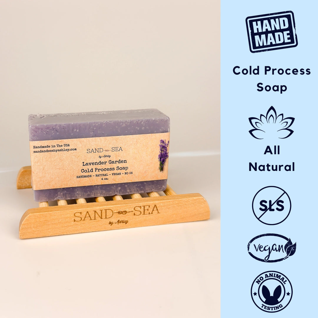 Bar of soap on a wooden stand with 'Sand & Sea' branding, next to icons indicating 'Hand Made', 'Cold Process Soap', 'All Natural', 'SLS Free', 'Vegan', and 'No Animal Testing'.