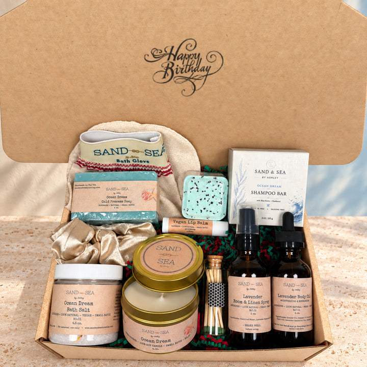 A curated Sand &amp; Sea gift box set displayed inside an open kraft cardboard box. The set includes bath products like a bath glove, bath bomb, shampoo bar, and green sea salts, along with skincare items such as a lip balm, a small wooden box, and two amber spray bottles.