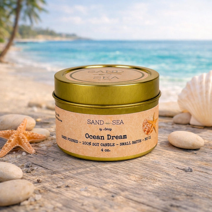 Candle in a tin labeled 'Sand of the Sea' on a beach setting with ocean and shells in the background.