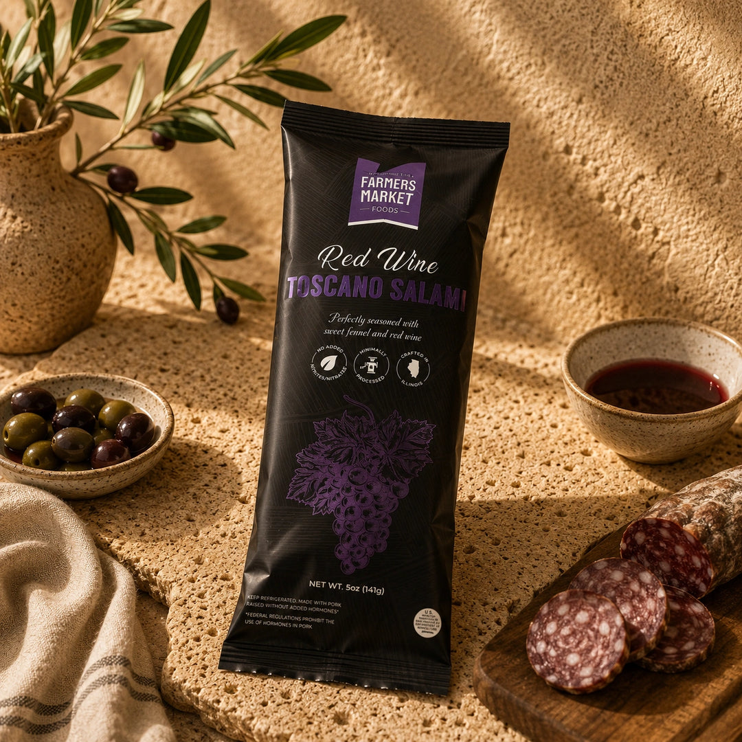 A package of Farmers Market Red Wine Toscano Salami stands upright on a textured stone surface, surrounded by olive branches, olives, a bowl of red wine, and sliced salami on a wooden board.