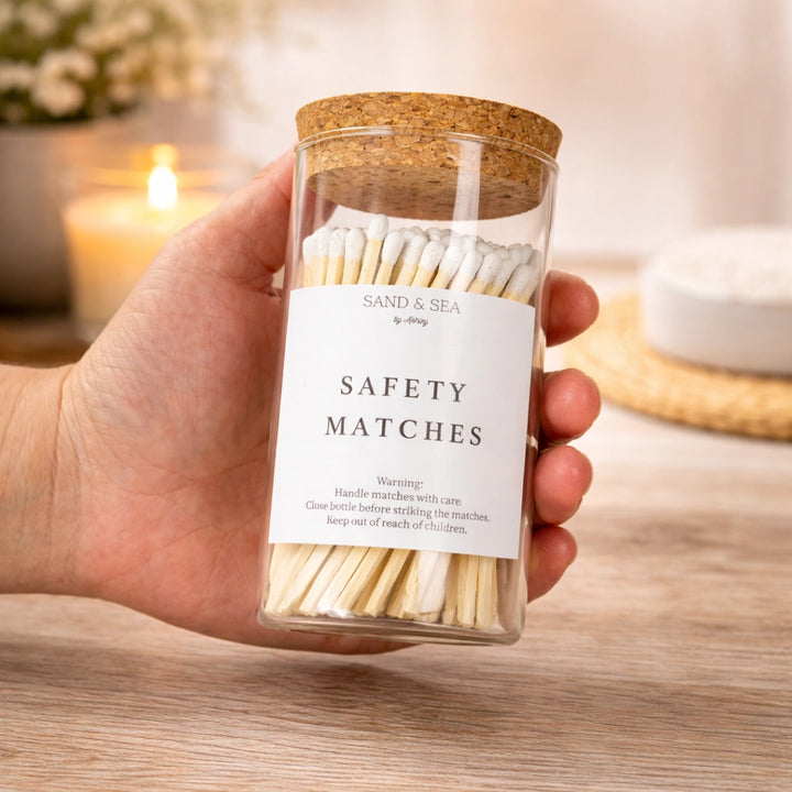 Hand holding a jar of safety matches labeled 'Sand & Sea' with a blurred candle in the background.