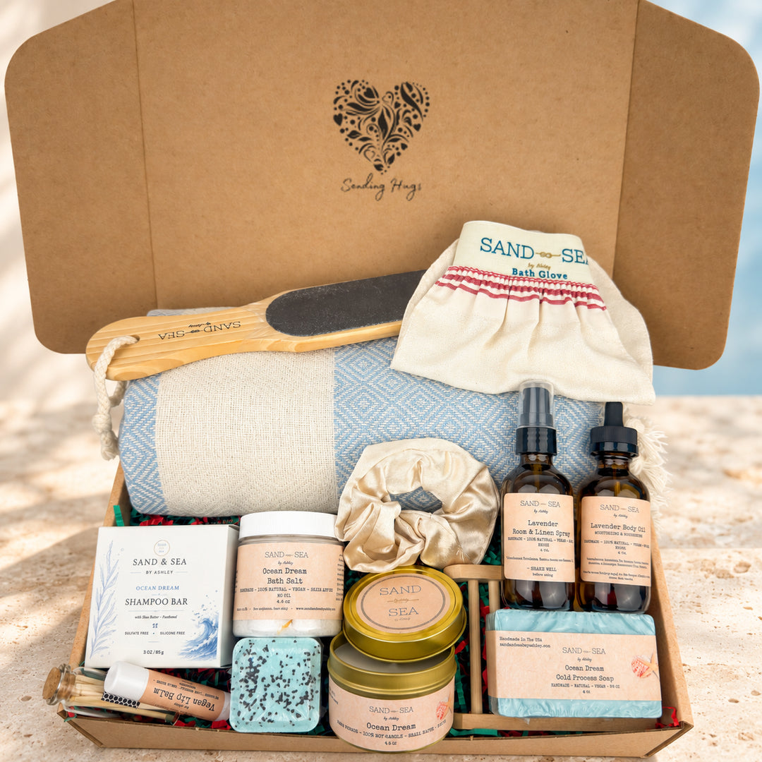 A curated self-care gift box from Sand & Sea Spa by Seeing Hugs, filled with spa items including a blue-and-white patterned towel, wooden hairbrush, cotton scrunchie, bath bomb, shampoo bar, tins, and dropper bottles, arranged in an open kraft cardboard box on a beach-like surface.