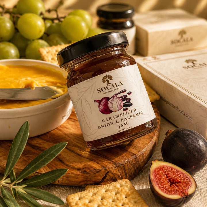A glass jar of Socala Caramelized Onion & Balsamic Jam is styled on a wooden board with crackers, green grapes, a bowl of yellow spread, olives, and a fig, alongside branded gift boxes.