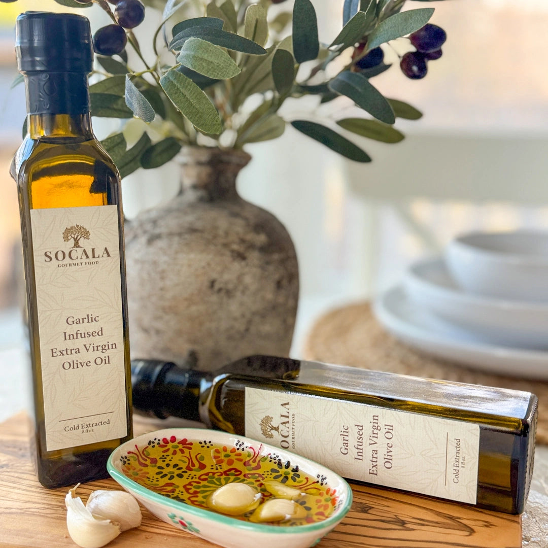 Two bottles of Socala Garlic Infused Extra Virgin Olive Oil on a wooden surface with a decorative bowl and olive branch.