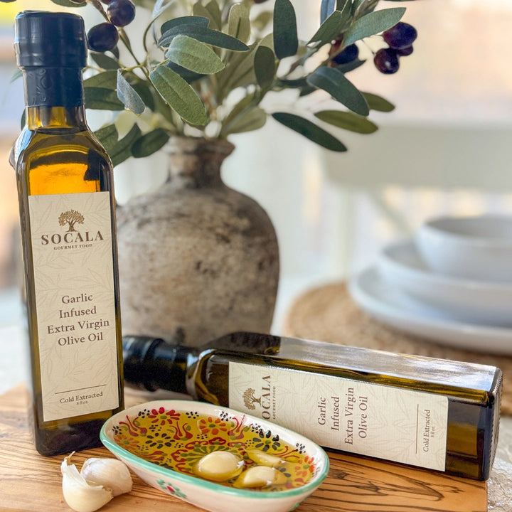 Two bottles of Socala Garlic Infused Extra Virgin Olive Oil on a wooden surface with a decorative bowl and olive branch.