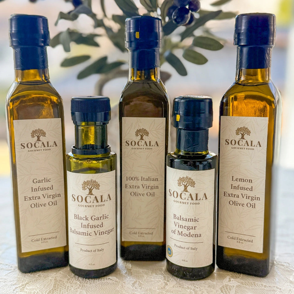 Collection of Socala olive oil and vinegar bottles on a textured surface with blurred greenery in the background.