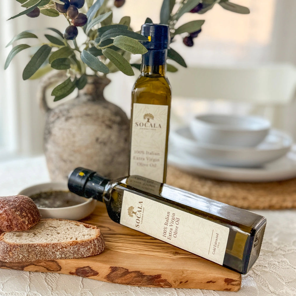 Two bottles of Socala olive oil on a wooden cutting board with bread and olive branches.