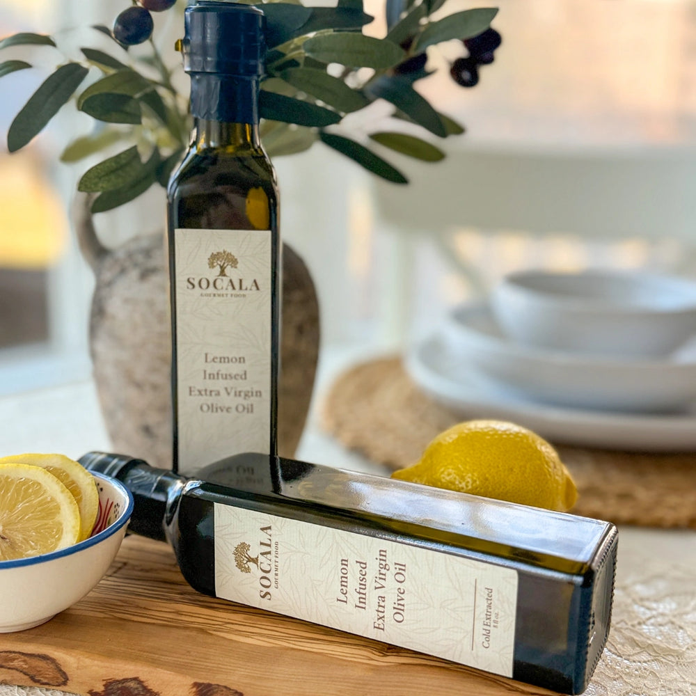 Two bottles of Socala Lemon Infused Extra Virgin Olive Oil on a wooden surface with lemons and olive branches.