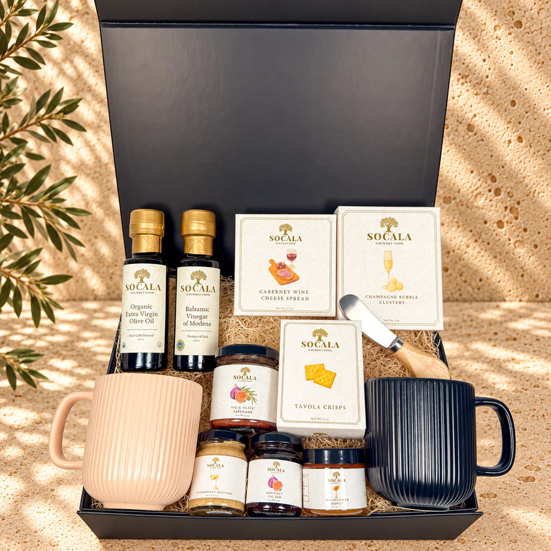 A curated gift basket set from Socala, presented in a black gift box on a sandy surface with dappled sunlight and shadows. The set includes two small bottles of vinegar, two boxes of Socala cheese and crackers, two jars of spreads, a wooden-handled cheese knife, a light pink ribbed ceramic mug, and a dark navy ribbed ceramic mug.