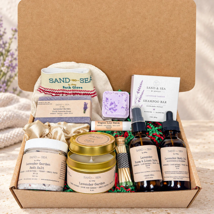 A Sand &amp; Sea bath and body gift box filled with multiple lavender-themed products including bath salts, a bath glove, shampoo bar, body spray, and jars, arranged in a kraft cardboard box with green paper filler.