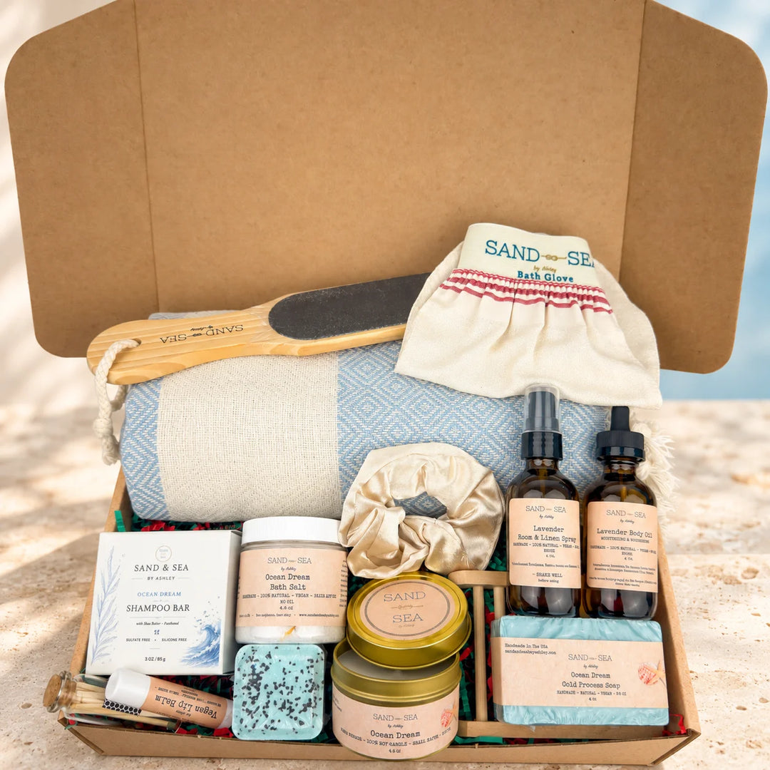 An open cardboard gift box filled with a spa-style bath set, including a blue patterned towel, wooden scrub brush, cream bath glove, shampoo bar, bath bomb, soap, and two amber bottles, photographed on a sandy beach with water in the background.