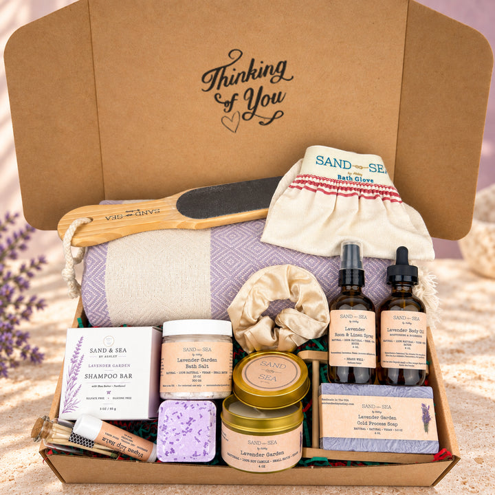 A curated bath and body gift box from Sand &amp; Sea, filled with lavender-themed products including a lavender shampoo bar, lavender body wash, lavender bath salts, lavender essential oil, a lavender scrubber, a lavender body soap bar, a lavender silk scrunchie, a lavender bath glove, and a lavender pillowcase, all arranged in a brown cardboard box with the lid printed with &