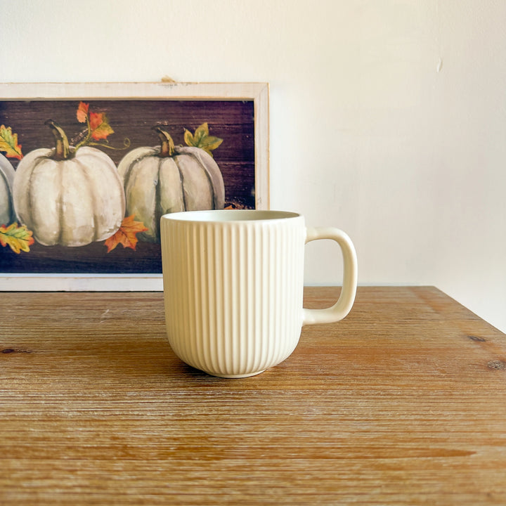 White ceramic mug on a wooden surface with a framed picture of pumpkins in the background.